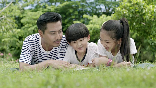 亚洲父亲和他的孩子们躺在草原上读故事书视频购买