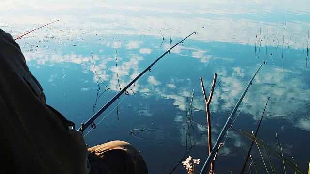 钓鱼杆在渔夫手中。湖岸边。平静而平静的水面上有云的倒影。美丽的自然风景。渔夫在钓鱼。鱼竿被抛入水中，等待鱼儿上钩视频素材