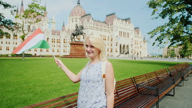 一名年轻的女游客在布达佩斯议会的背景下与匈牙利国旗摆姿势。在欧洲旅游视频下载