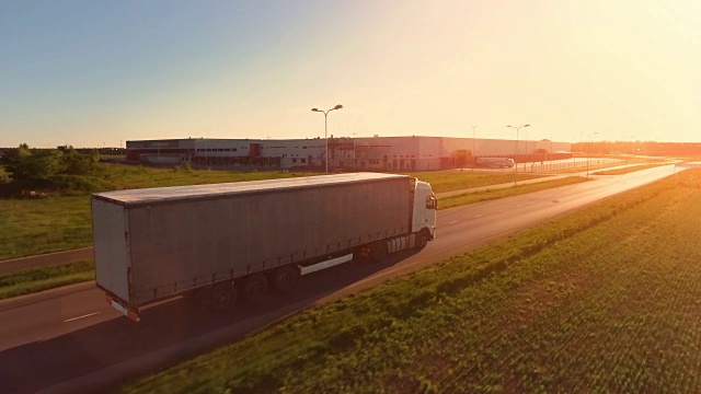 在高速公路上移动的白色半挂车的鸟瞰图。在背景中可以看到仓库和工业装载建筑。日落。视频素材