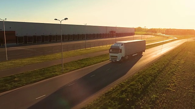 在高速公路上移动的白色半挂车的鸟瞰图。在后台仓库和农村地区，太阳正在下山。视频素材
