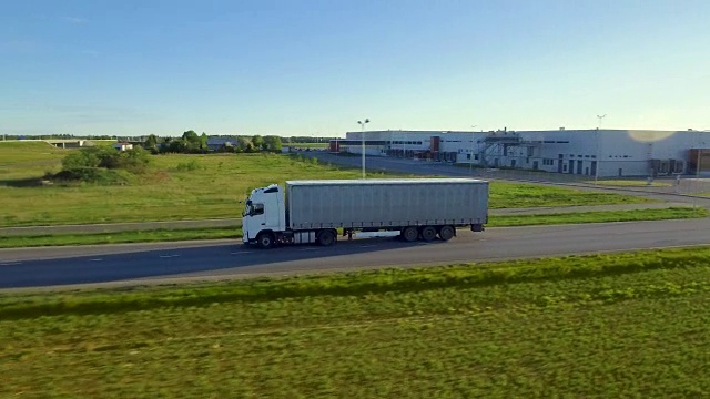 白色半挂车与货运拖车在高速公路上移动的鸟瞰图。背景是仓库和农村，美丽的晴天。视频素材