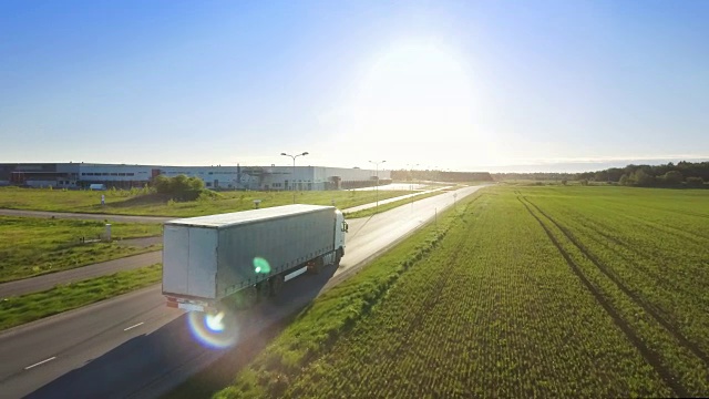 鸟瞰图的白色卡车与货物半挂车连接在高速公路上的驱动器。在背景中可以看到仓库和工业装载建筑。日落。视频素材
