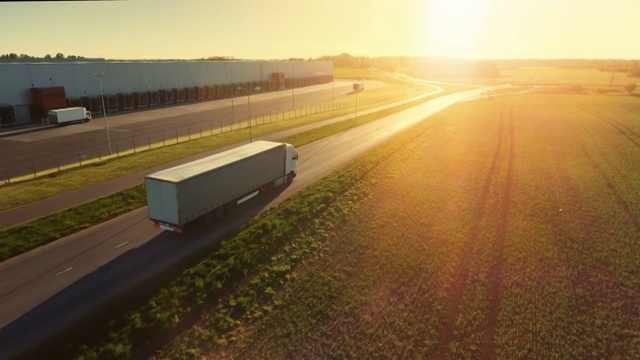 空中跟踪拍摄的白色半挂车与货运拖车通过工业仓库，农村地区。阳光普照，天空湛蓝。视频素材