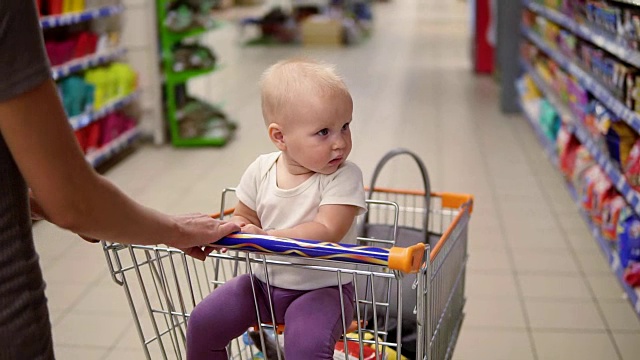 一个可爱的小婴儿坐在超市的购物车里四处张望。母亲的手推着手推车在不同的部门之间向前移动。全家带着小孩购物视频素材