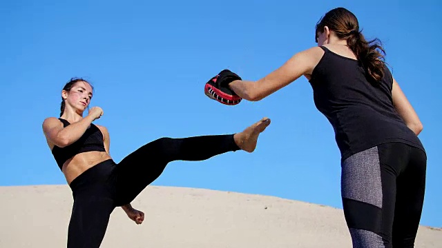 两个运动的年轻女子穿着黑色的健身服，在空旷的海滩上，在蓝天下，在炎热的太阳下，锻炼踢腿，训练打架。慢动作视频下载