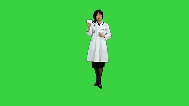 迷人的女医生穿着实验室大褂，带着听诊器在绿屏上和手机通话视频下载