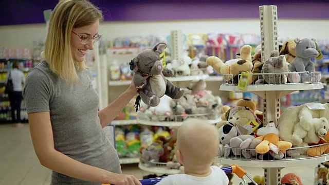 微笑的母亲在超市儿童玩具区给她的孩子看玩具大象，而她可爱的孩子坐在购物车里。家庭购物视频购买