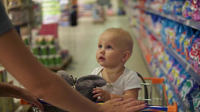 一个小婴儿抱着玩具坐在超市的购物车里，而她的妈妈推着购物车在挑选商品。全家带着小孩购物视频素材
