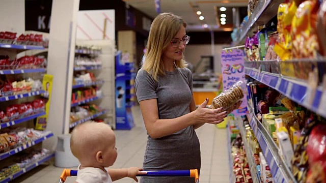 年轻的母亲正在阅读饼干包装上的产品内容，而她的孩子正坐在超市的购物车里视频下载