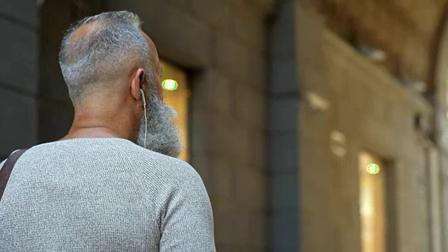 一个戴着耳机的灰白头发的男人在城市里散步视频素材