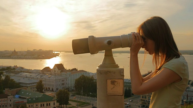 年轻女子透过旅游望远镜，在夕阳下探索城市视频素材