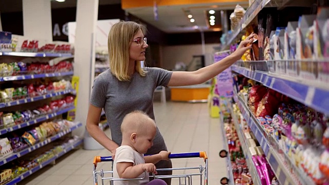 一位年轻漂亮的妈妈正在超市挑选糖果和蛋糕，而她的小宝宝正坐在购物车里。家庭购物时间视频下载