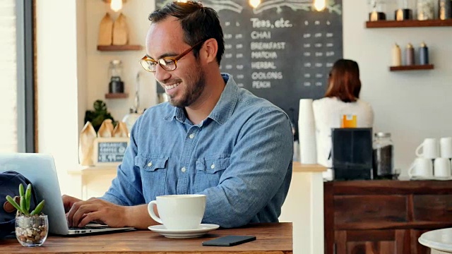 一个快乐的西班牙裔年轻人微笑着在咖啡店里使用电脑视频下载