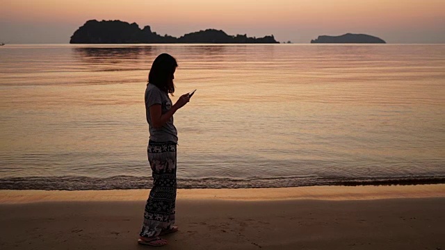 年轻的亚洲女人在日出的海滩上使用智能手机。视频素材