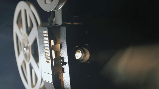 一个老式的古董超级8毫米电影放映机的正面视图，投射一束光在一个黑暗的房间旁边的一堆散开的电影卷轴视频素材