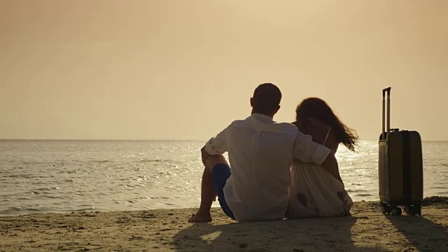抵达度假。一对年轻夫妇坐在他们旅行袋旁边的沙滩上。欣赏海面上的日落视频素材