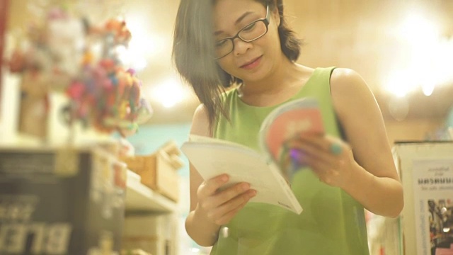 亚洲妇女微笑，而阅读一本书在书店对书架的背景。视频下载