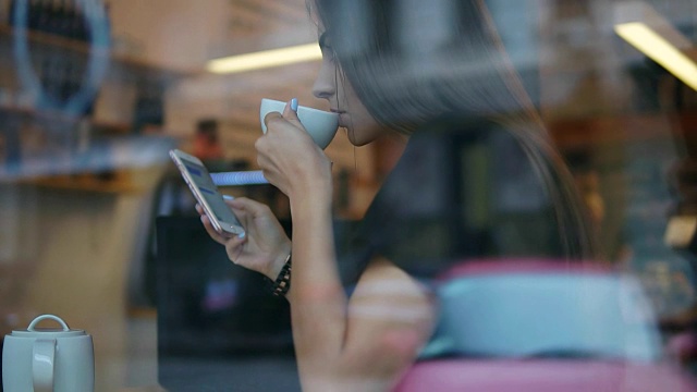从大街上看，一个留着长发的年轻女子正在用手机发短信，喝着咖啡店里的白色杯子里的茶或咖啡视频素材