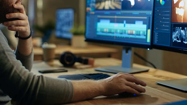一名男性摄像师在他的个人电脑上用两个显示器编辑和剪辑镜头和声音。他的办公室是现代创意Loft工作室。视频素材