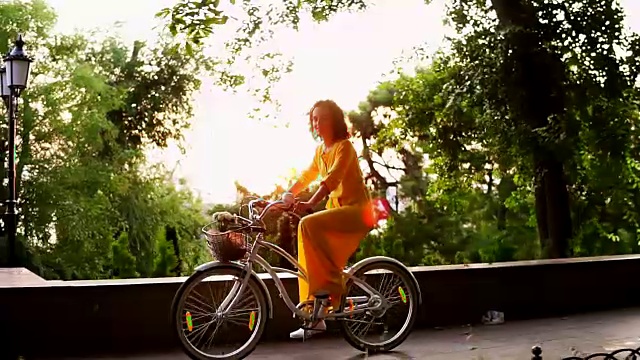 一个穿着黄色长裙的漂亮女人骑着一辆城市自行车，里面有一个篮子和鲜花。镜头光晕。替身。Slowmotion视频素材