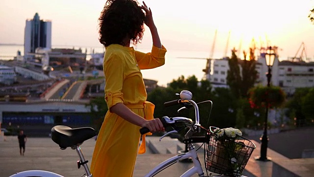 迷人的黑发女子穿着黄色长裙享受黎明站在她的城市自行车的车把与鲜花在其篮子享受清晨。镜头光晕，美丽的城市景观视频素材