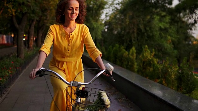 穿着黄色长裙的美丽女子骑着一辆城市自行车，带着篮子和鲜花在城市公园里看着镜头微笑着。替身。Slowmotion视频素材