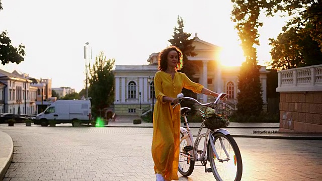 身着黄色长裙的黑发女子微笑着走在黎明的路上，手里握着城市自行车的车把，花篮里放着鲜花。镜头光晕，美丽的城市景观视频素材