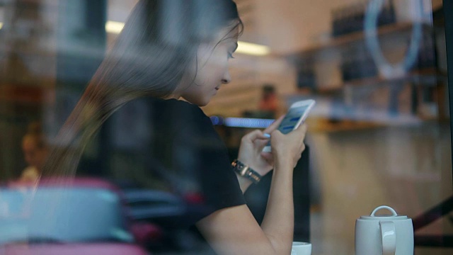 从外面看，一个长头发的年轻女子在咖啡店里用手机发短信。木桌上放着白色的茶杯、茶托和茶壶视频素材