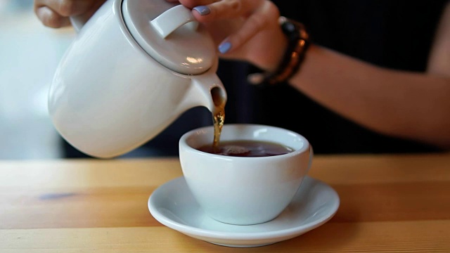 近距离观看女性的手倒热茶从茶壶在咖啡馆的木桌上的白瓷杯视频素材