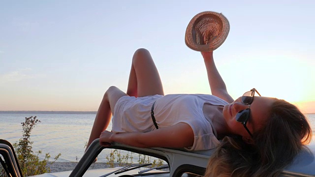 快乐的旅程，放松女孩在机器在海边，夏天的旅行，年轻女子躺在车顶在海边的河流，女性躺在汽车上视频素材