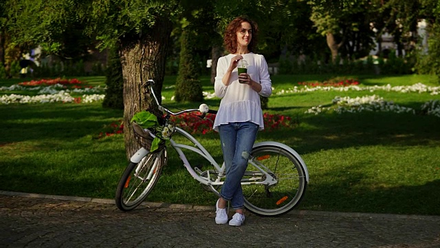 美丽的女人站在城市公园附近，在她的城市自行车的花篮和喝绿色排毒奶昔。健康的生活方式视频素材