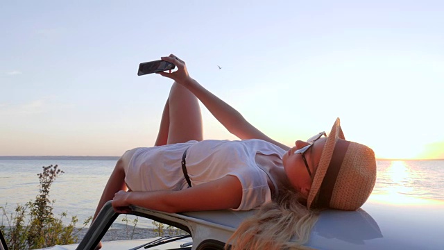 手机在快乐的旅程上，可爱的女孩把手机放进手躺在车顶，图片手机做智能手机在怀里的女孩视频素材