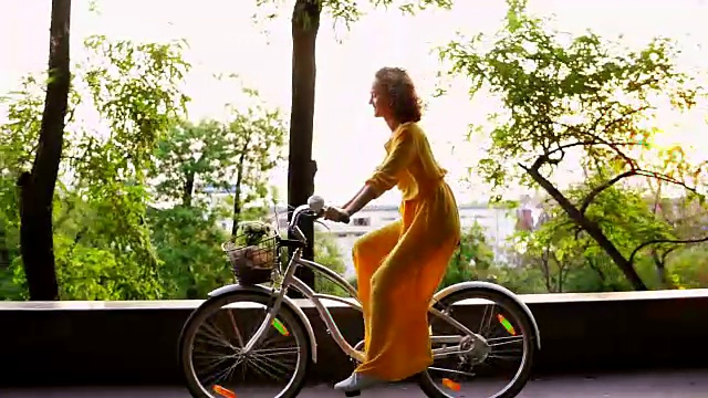 身着黄色长裙的褐发女人带着篮子和鲜花在黎明时分骑着一辆城市自行车，面带微笑。镜头光晕。替身。Slowmotion视频素材