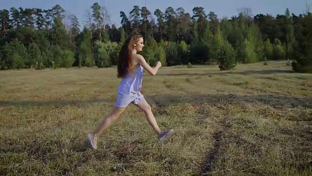 夏天的傍晚，一个快乐的年轻女子带着棕色的长卷发跑过松林附近的田野视频素材