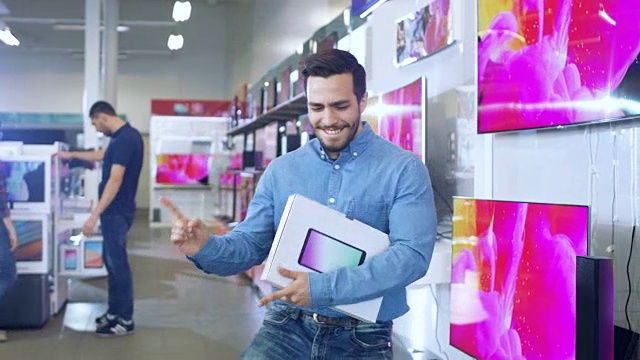 一名年轻人在电子商店购买了最新款的平板电脑，并高兴地与盒子跳舞。明亮和现代商店有所有最新的设备在商店。视频下载