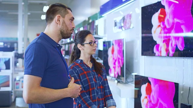 在电子商店专业顾问提供专家建议给一个年轻女子谁寻找一个新的4K超高清电视购买。在这个明亮、现代的商店里，所有最新的电视型号都挂在墙上。视频素材