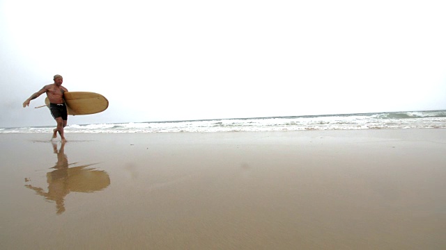 一个成熟的男性冲浪者在玻璃般光滑的沙滩上奔跑。视频素材
