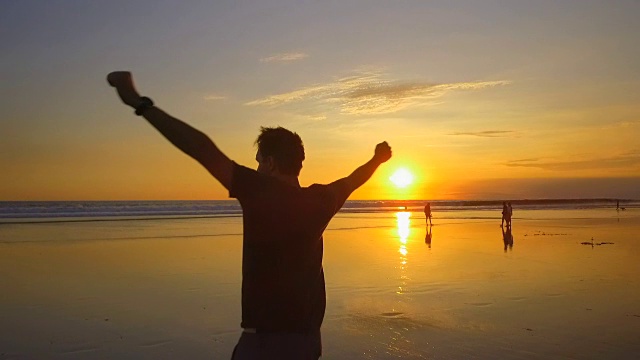 慢镜头:日落时分，一名快乐的游客高举双手，在海滩上旋转视频下载