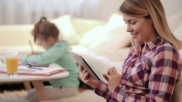 快乐的母亲在家里使用数字平板电脑，而她的女儿在背景上色。视频素材