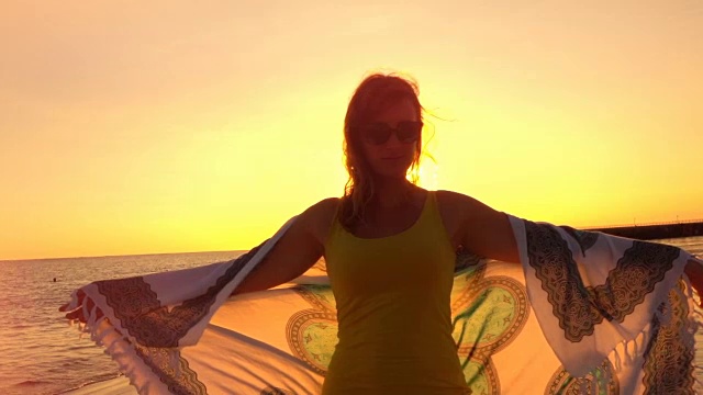 慢镜头:夕阳西下，美丽的女孩手拿飘动的围巾站在沙滩上视频素材