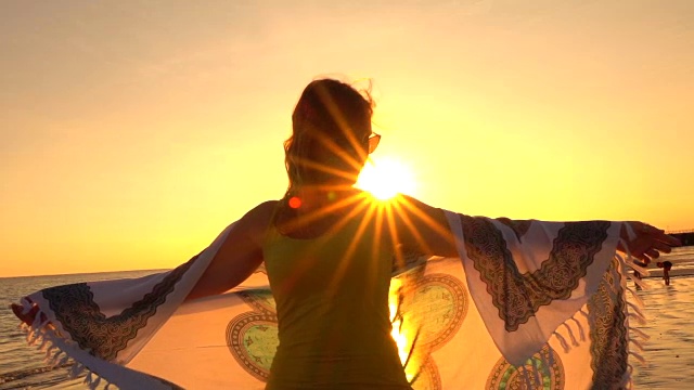 慢镜头落日时分，迷人的女子披着飘动的围巾站在沙滩上视频素材