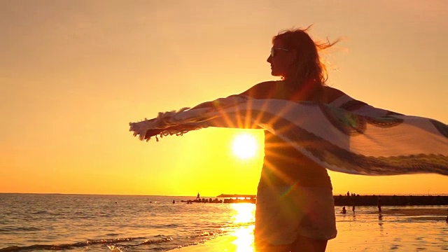 慢镜头:日落时分，戴着太阳镜和飘动的围巾的女孩在海滩上旋转视频购买