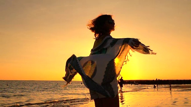 慢镜头:金色的夕阳下，一个女人围着飘动的围巾在沙滩上旋转视频素材