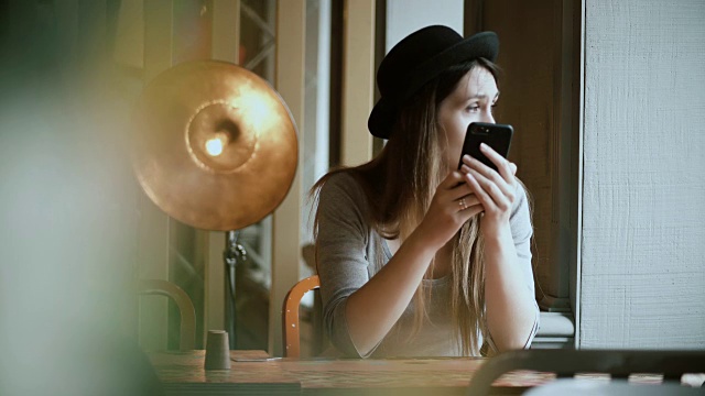 年轻美丽的女人戴着帽子，拿着智能手机，在咖啡馆使用无线技术的肖像视频下载