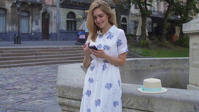 年轻女孩坐在城市的喷泉上玩智能手机。快乐的年轻女子微笑着使用iPhone智能手机的摄像头。旅游，instagram，现代科技。视频素材