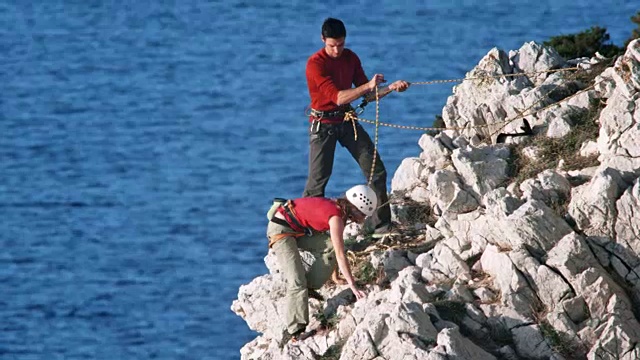 一名男子祝贺他的女性攀岩伙伴，因为她到达了海边一个参差不齐的岩石的顶端视频素材
