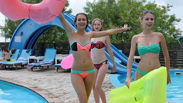 女孩们穿着比基尼在泳池边的木栈桥上走着，带着粉色的橡胶圈，女朋友们穿着泳衣一起游泳视频素材