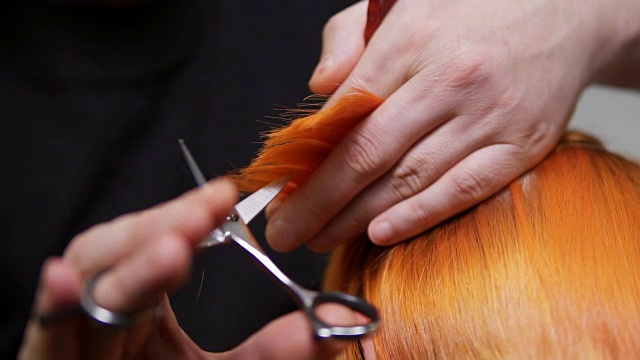 近距离观看红发女人的头发正在被一个专业的发型师在美容院剪。男性手握发丝，用剪刀和梳子剪。Slowmotion拍摄视频下载