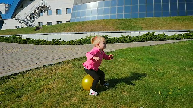 慢镜头:三岁女孩在草坪上的一个橡皮球上跳跃。视频下载